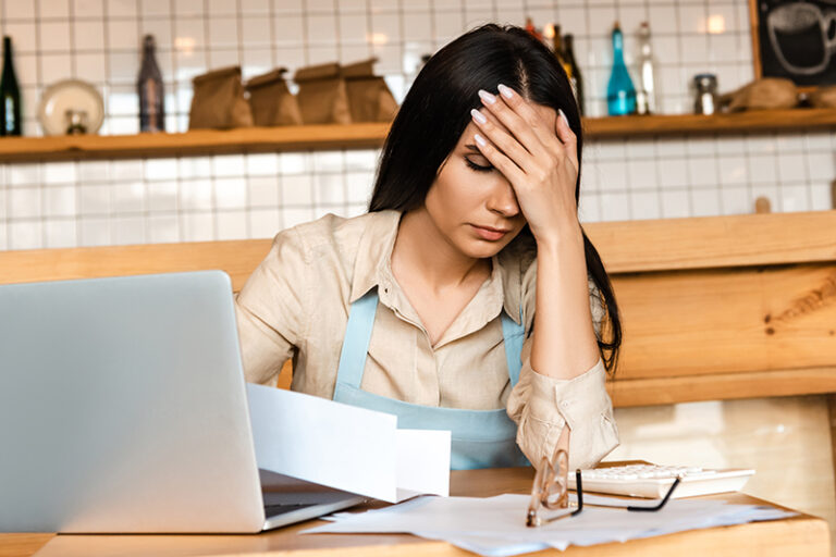 Upset cafe owner covering face near laptop, documents and glasses at table